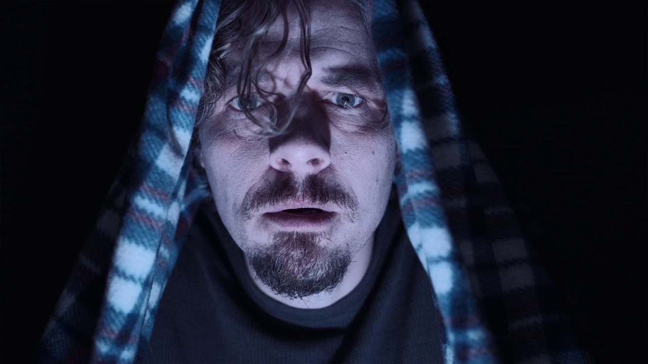 A startled man peers out beneath a checked blanket in Spoiling You