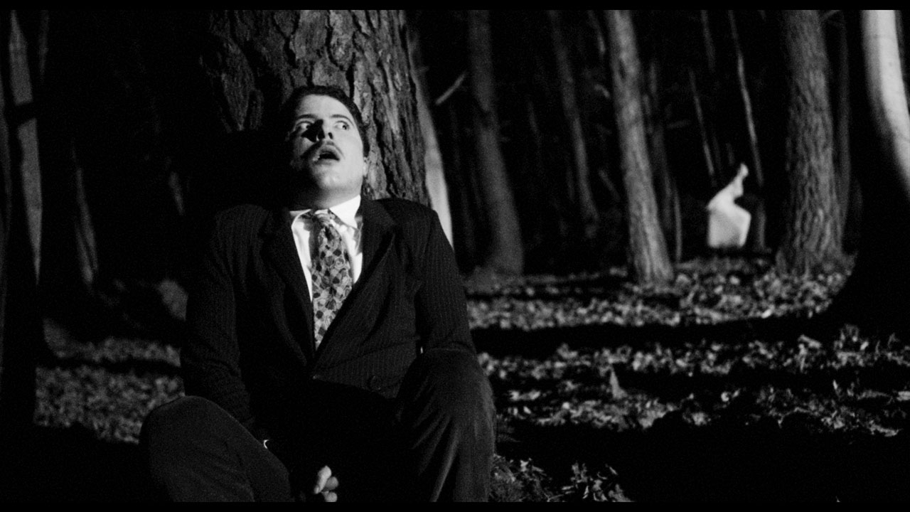 A frightened man sits against a tree in a dark woodland in Black Chariot